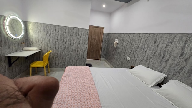 A hotel room with a bed, a bedside table, a chair, a mirror, and a door. - SRM INN (Ayodhya)