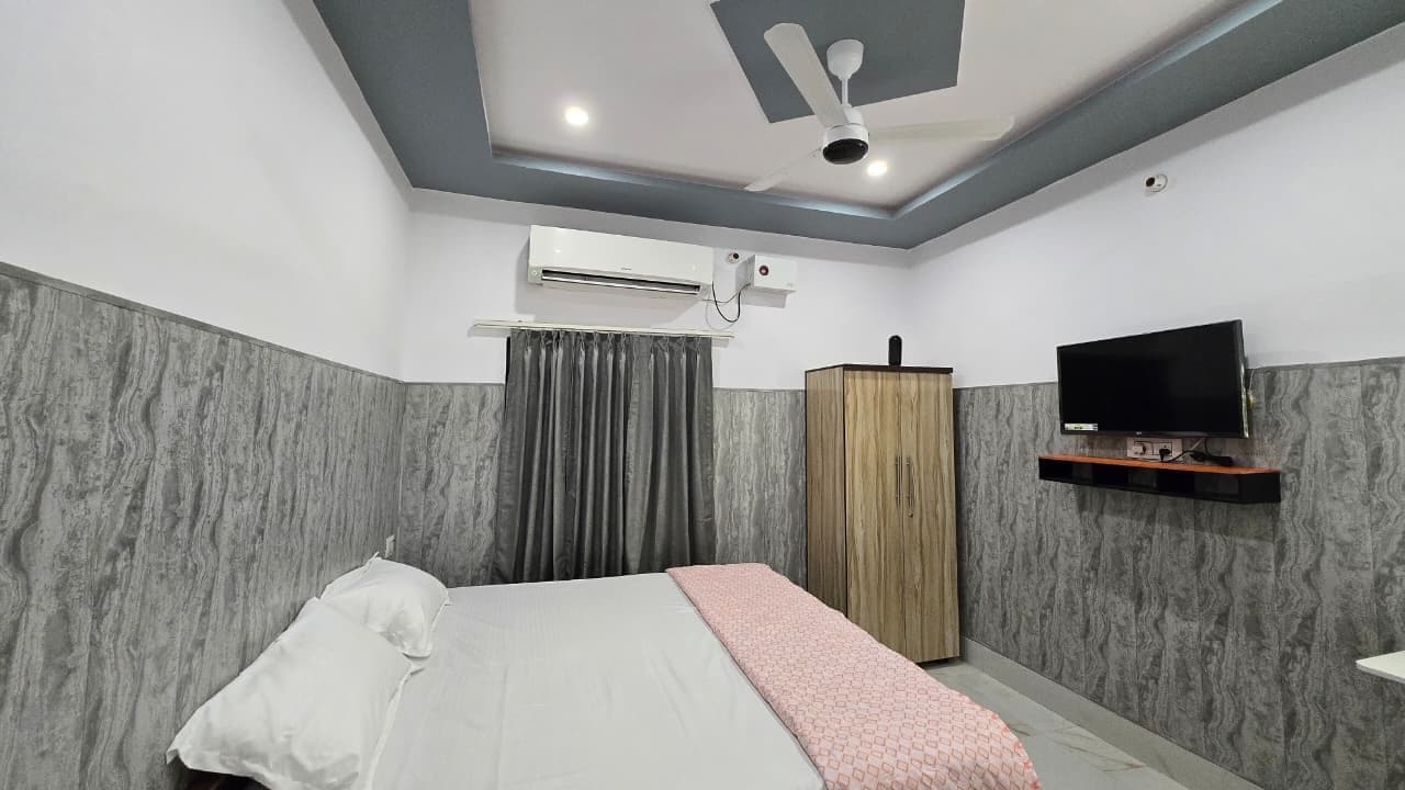 Une chambre à coucher avec un lit, un appareil de climatisation, un téléviseur posé sur une étagère en bois et un ventilateur de plafond.