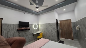 A hotel room with a bed, a desk with a chair, a television, a ceiling fan, and a door. - SRM INN (Ayodhya)
