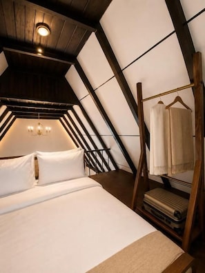 Une chambre avec un plafond en pente, un lit recouvert de draps blancs, un luminaire suspendu et une armoire en bois dotée d’un miroir.