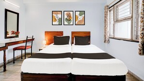 Ett hotellrum med en säng, ett skrivbord, en stol och ett fönster med gardiner. På väggen ovanför sängen hänger tre inramade konstverk.