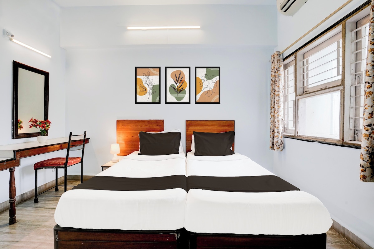 Quarto de hotel com cama, escrivaninha, cadeira e janela com cortinas. Três quadros emoldurados estão pendurados na parede acima da cama.