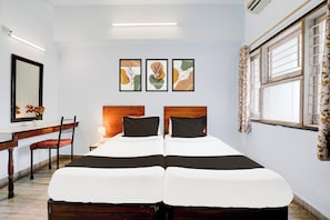Quarto de hotel com cama, escrivaninha, cadeira e janela com cortinas. Três quadros emoldurados estão pendurados na parede acima da cama.