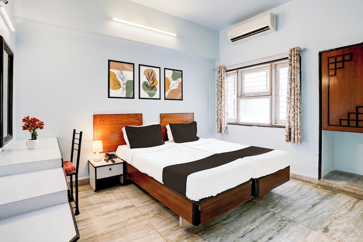 Quarto de hotel com cama, mesa de cabeceira, luminária, cadeira e janela com cortinas.