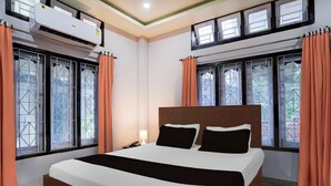 A hotel room with a large bed, two windows with curtains, an air conditioning unit, and a nightstand with a lamp. - Collection O Guwahati Airport (Guwahati)