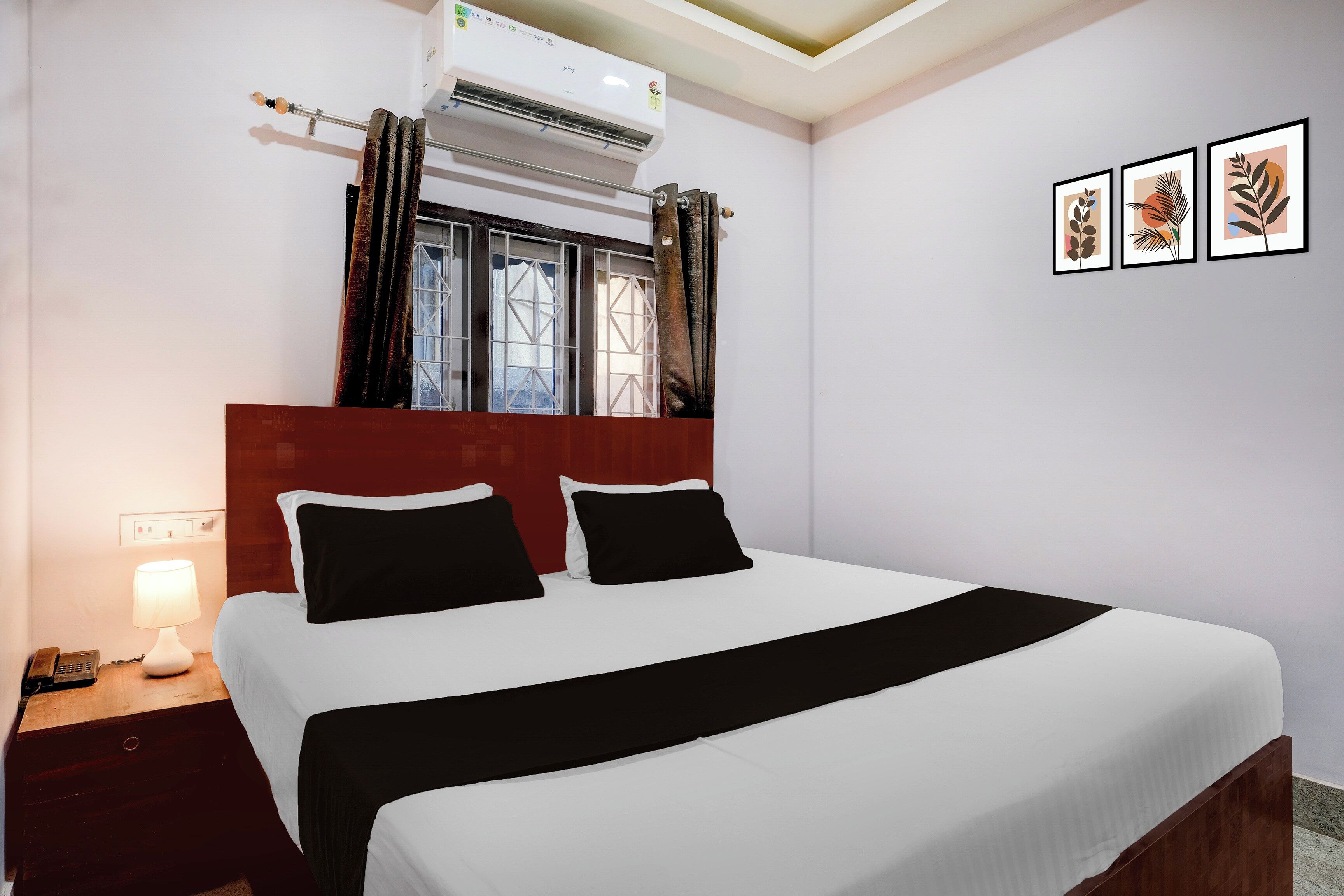 A hotel room with a bed, a nightstand, a lamp, an air conditioner, a window with curtains, and wall art.
