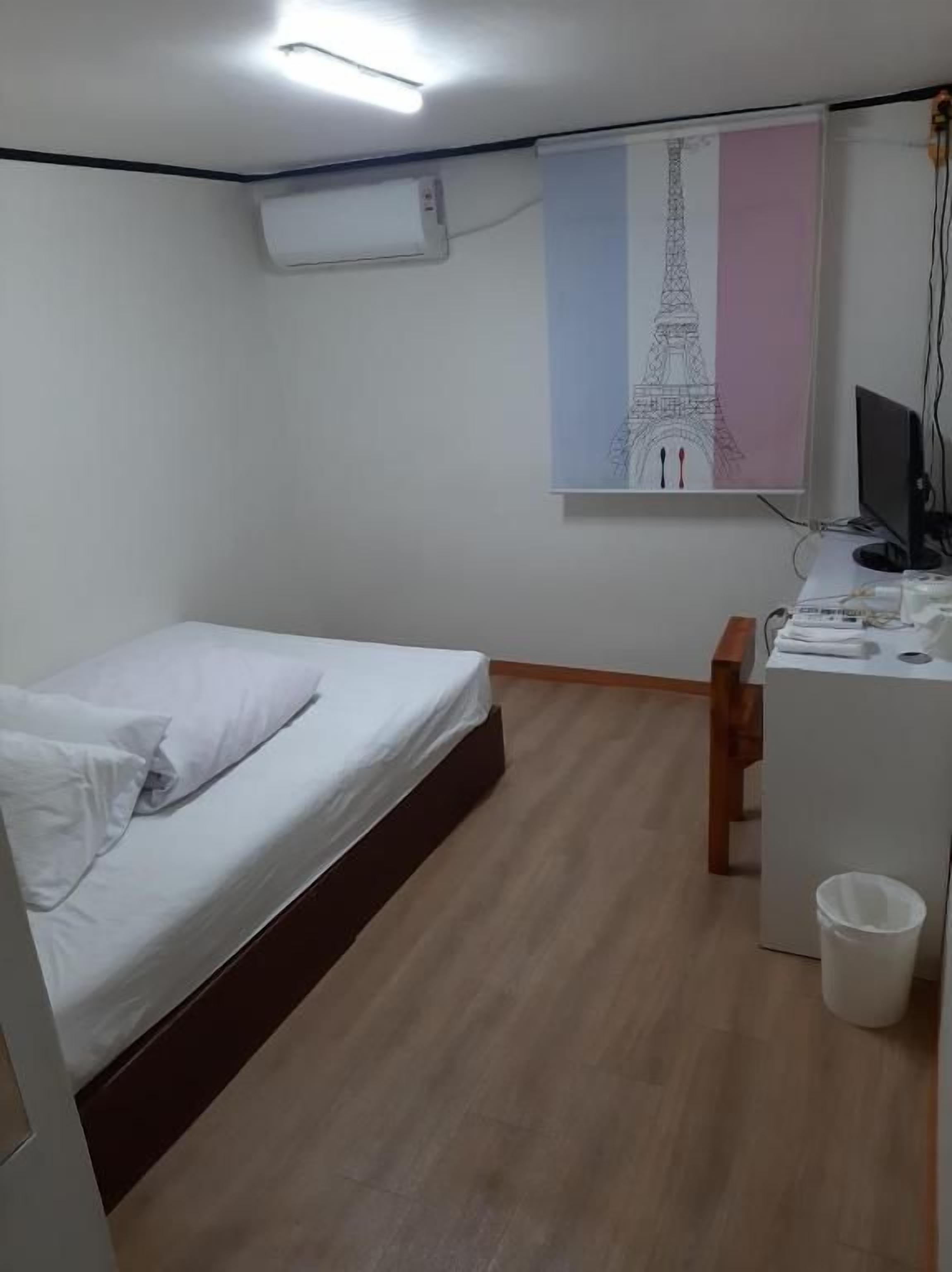 A bedroom with a bed, a desk, a chair, a computer, and an air conditioning unit.