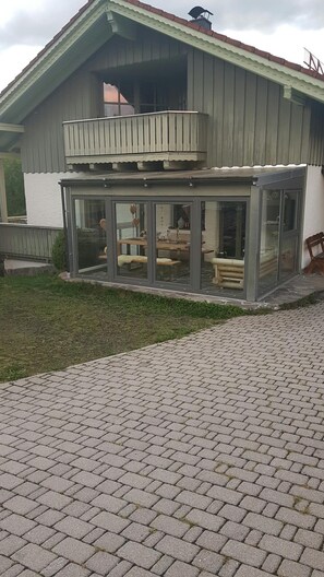 A modern house with a glass-enclosed patio area, a wooden deck, and a brick driveway. - Cozy vacation rental on the Guldensteig trail in the heart of the Bavarian Forest National Park (Neuschönau)
