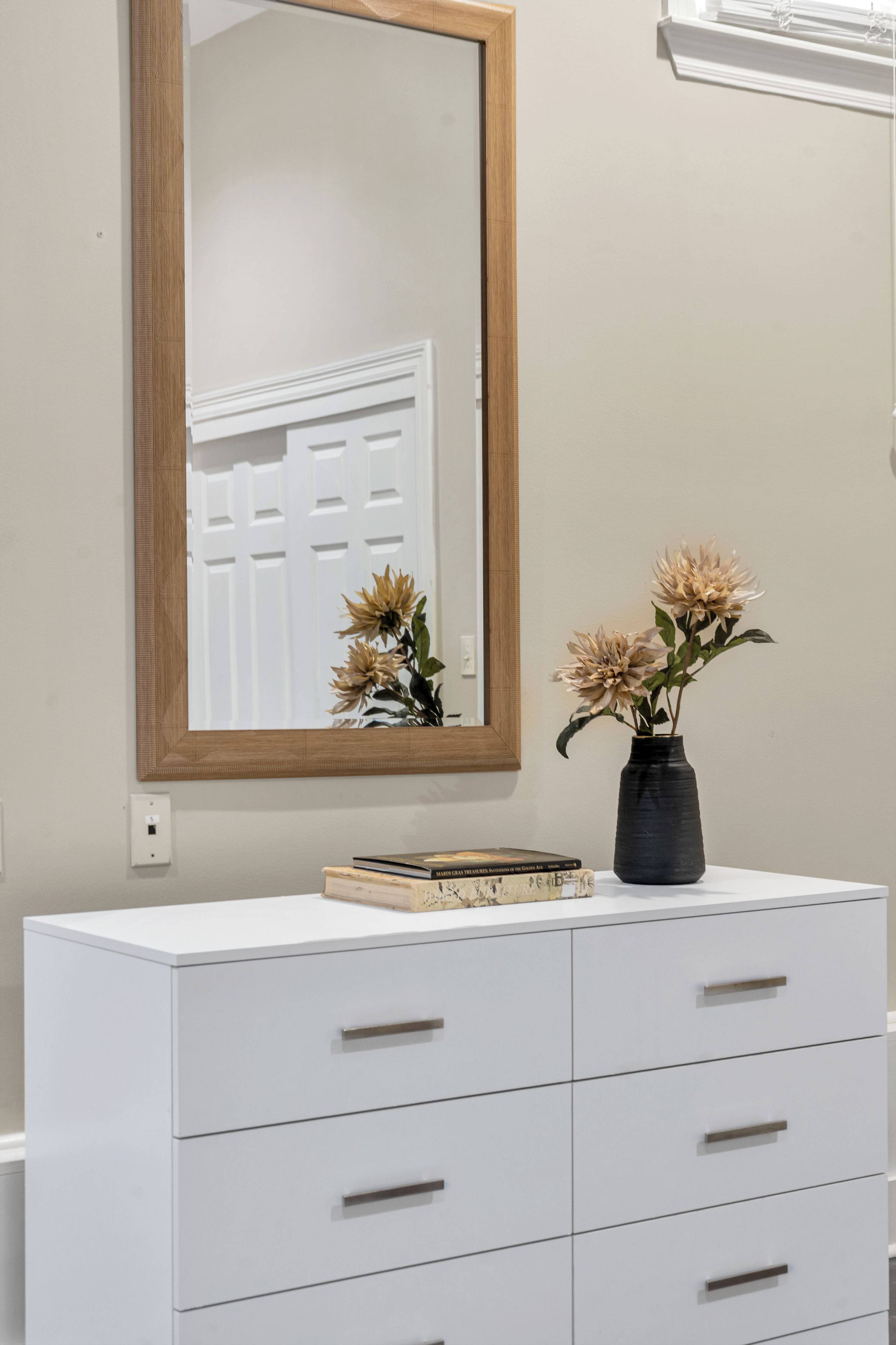 A white dresser with drawers, a mirror with a wooden frame, and a vase with dried flowers.