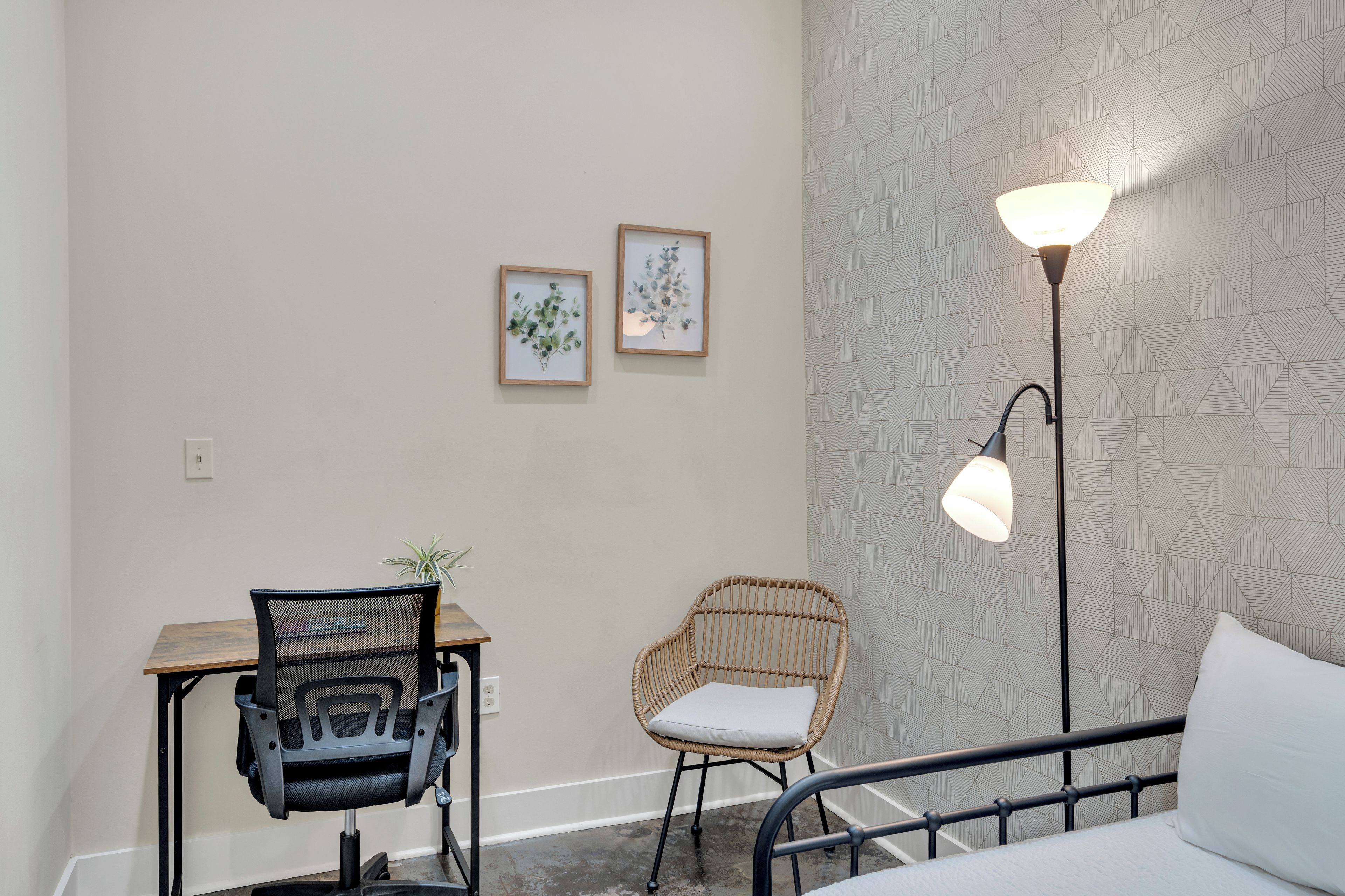 A room with a bed, a desk with a chair, a standing lamp, and two framed botanical prints on the wall.