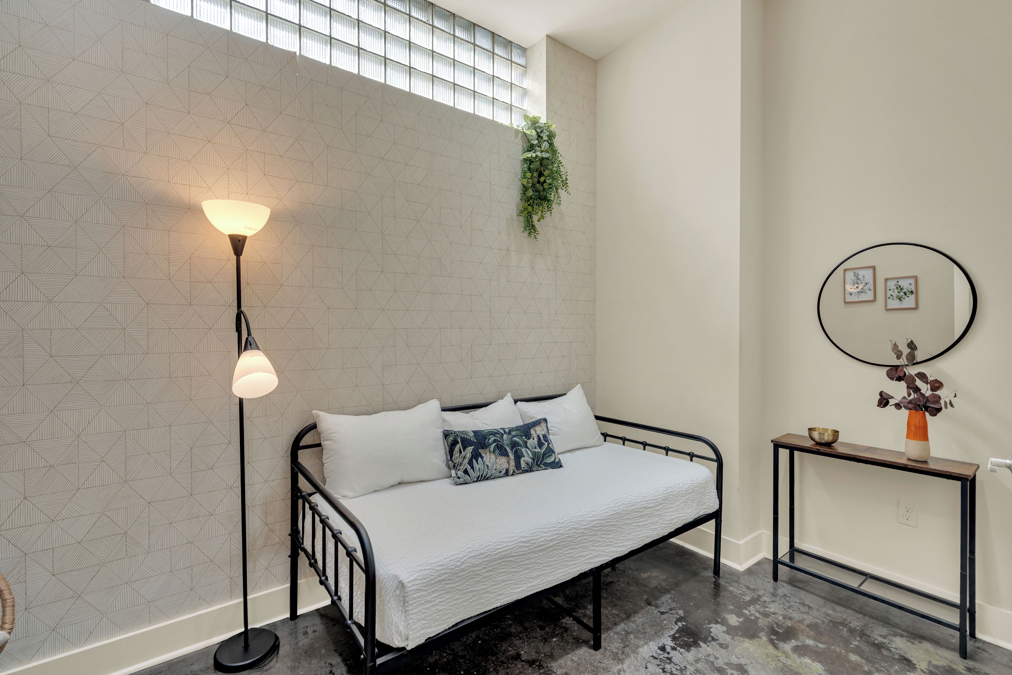 A bedroom with a bed, a floor lamp, a small table with a vase, and a wall with geometric patterns.