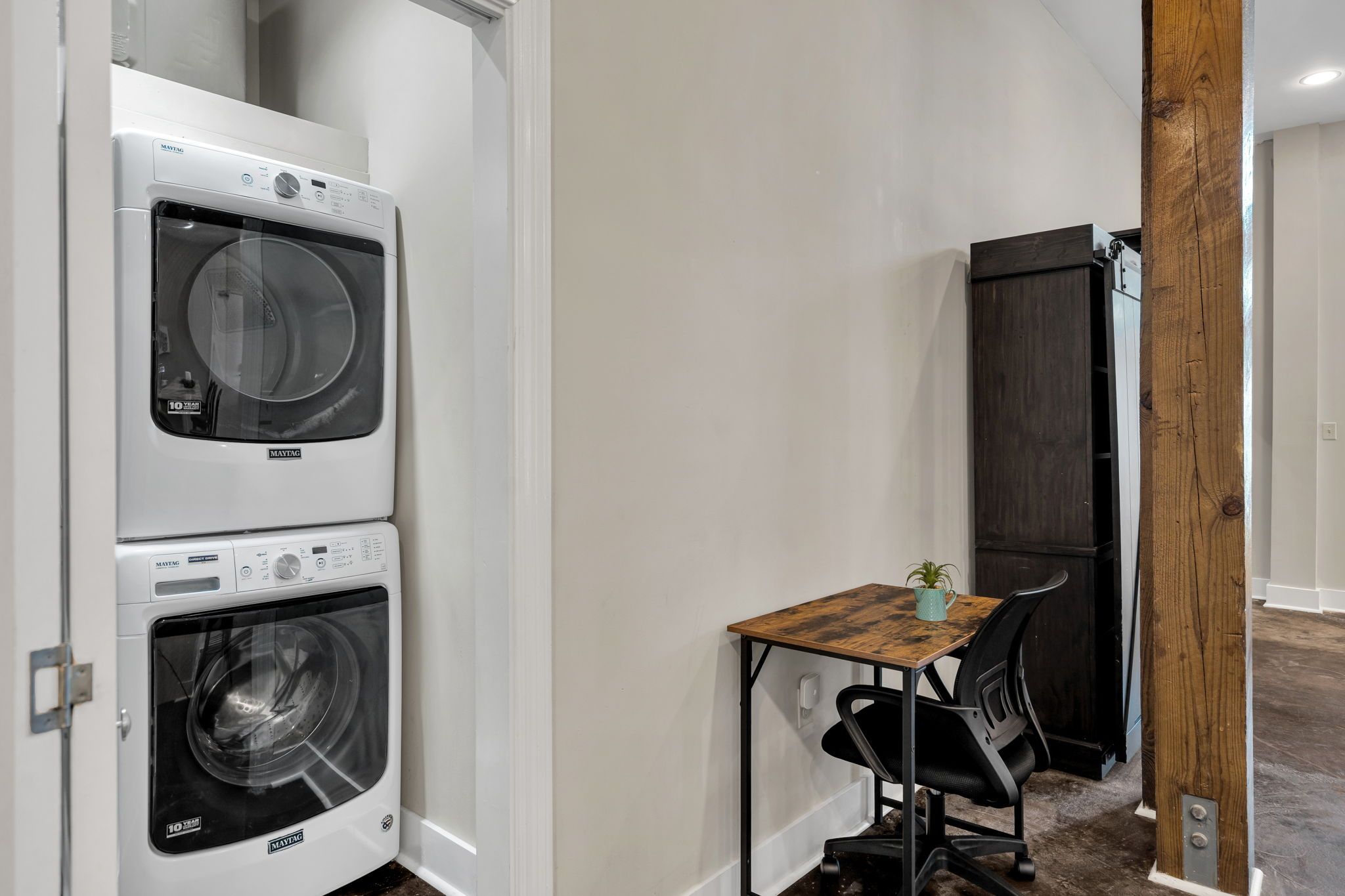 A laundry area with two stacked washing machines, a small table with a chair, and a wooden pillar.