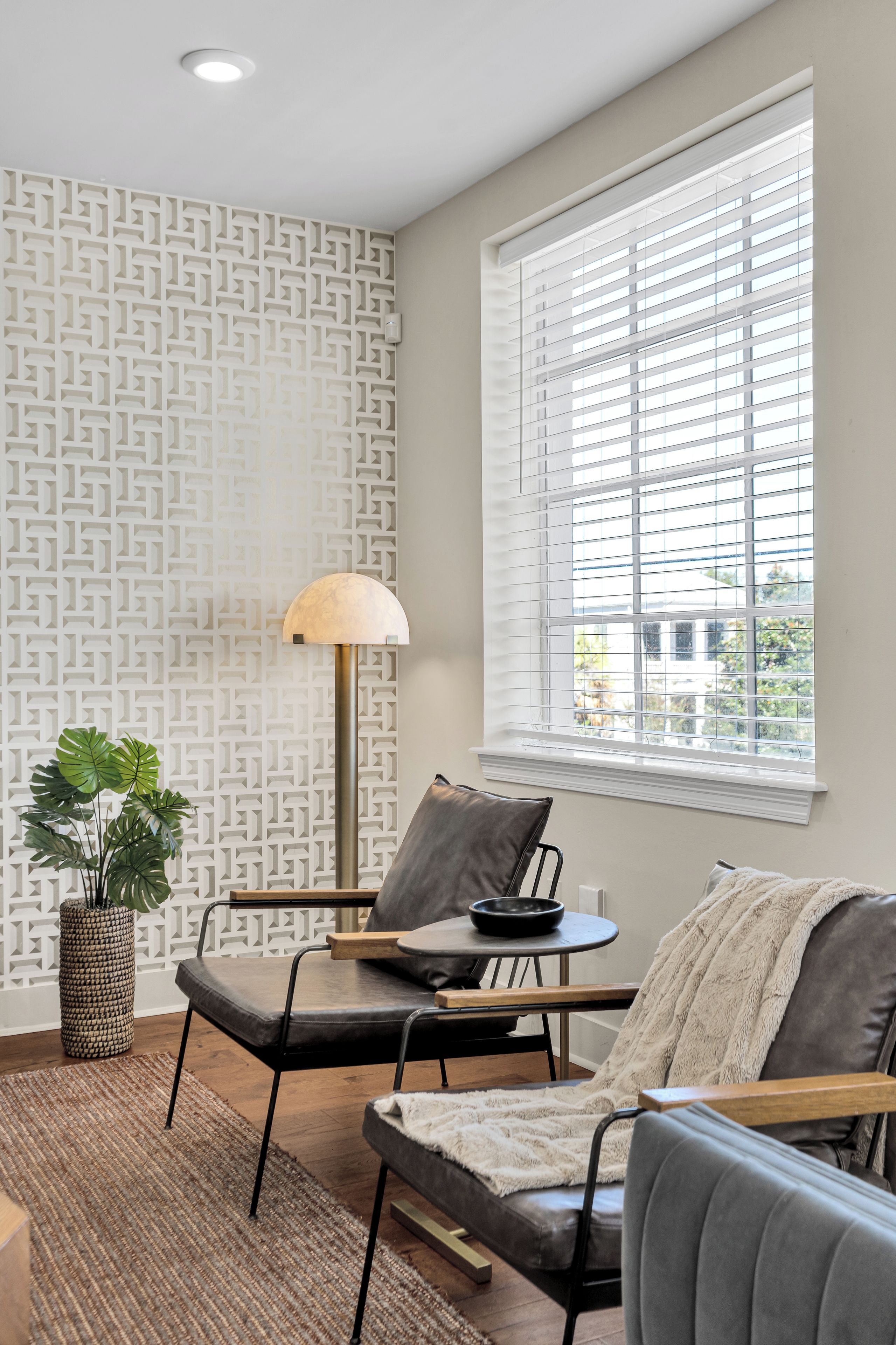 A modern living room with a patterned wall, a large window with blinds, a floor lamp, a sofa, and a side table with a bowl.