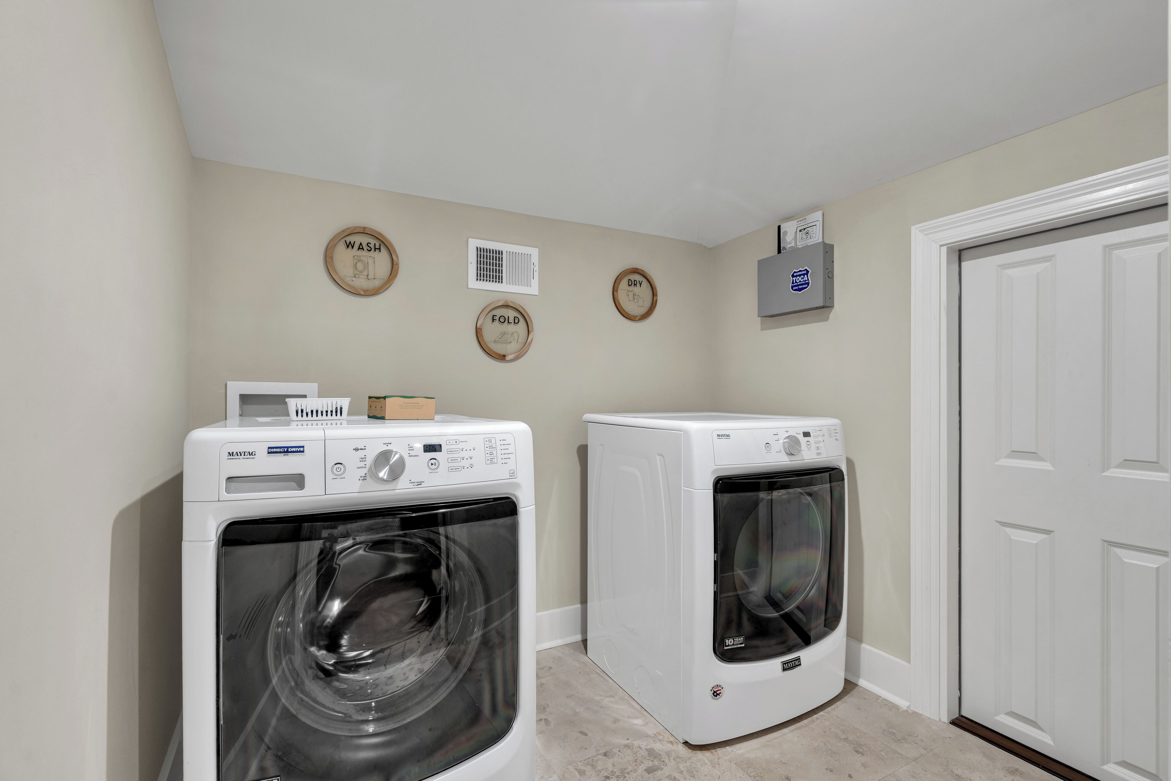 A laundry room with two Maytag washers and dryers, a wall-mounted air vent, and three wooden signs indicating 'WASH,' 'DRY,' and 'FOLD.'