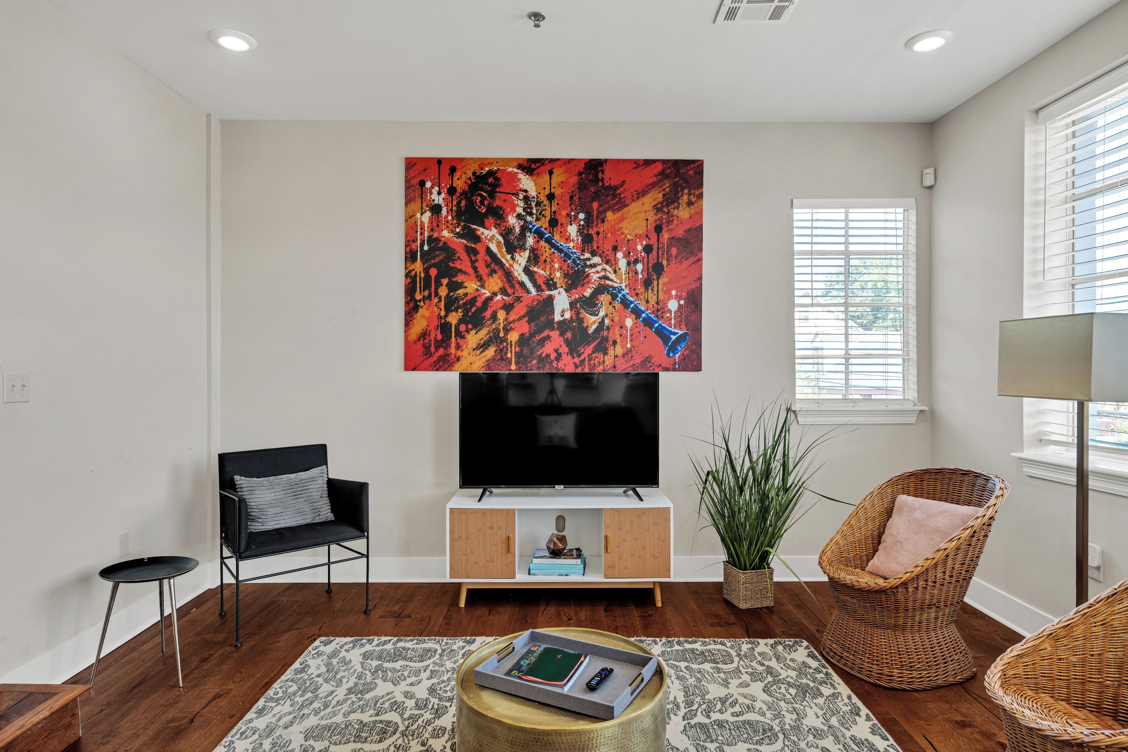 A modern living room with a flat-screen TV, a black armchair, a wicker chair, a potted plant, and a coffee table with books and remote controls.