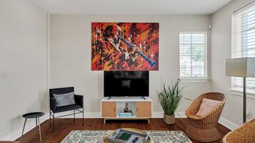 A modern living room with a flat-screen TV, a black armchair, a wicker chair, a potted plant, and a coffee table with books and remote controls.