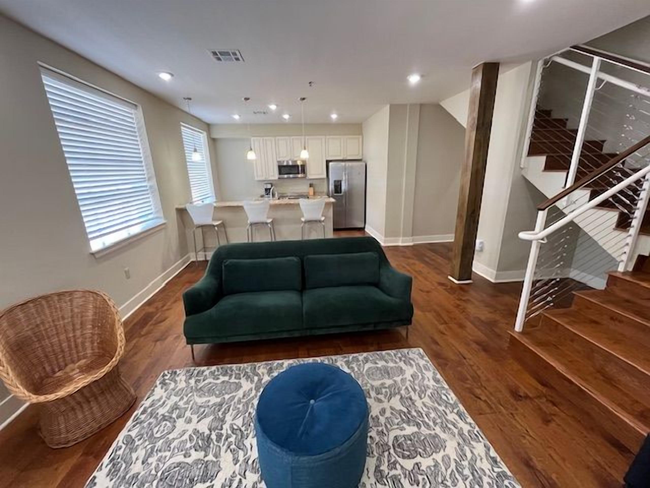 A modern living room with a green sofa, a patterned rug, and a wicker chair. A staircase is visible to the right.
