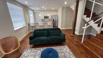 A modern living room with a green sofa, a patterned rug, and a wicker chair. A staircase is visible to the right.