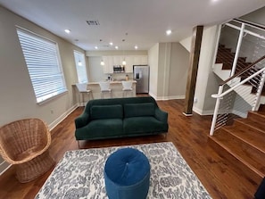 A modern living room with a green sofa, a patterned rug, and a wicker chair. A staircase is visible to the right. - Stylish 2BR condo near Frenchmen St (New Orleans)