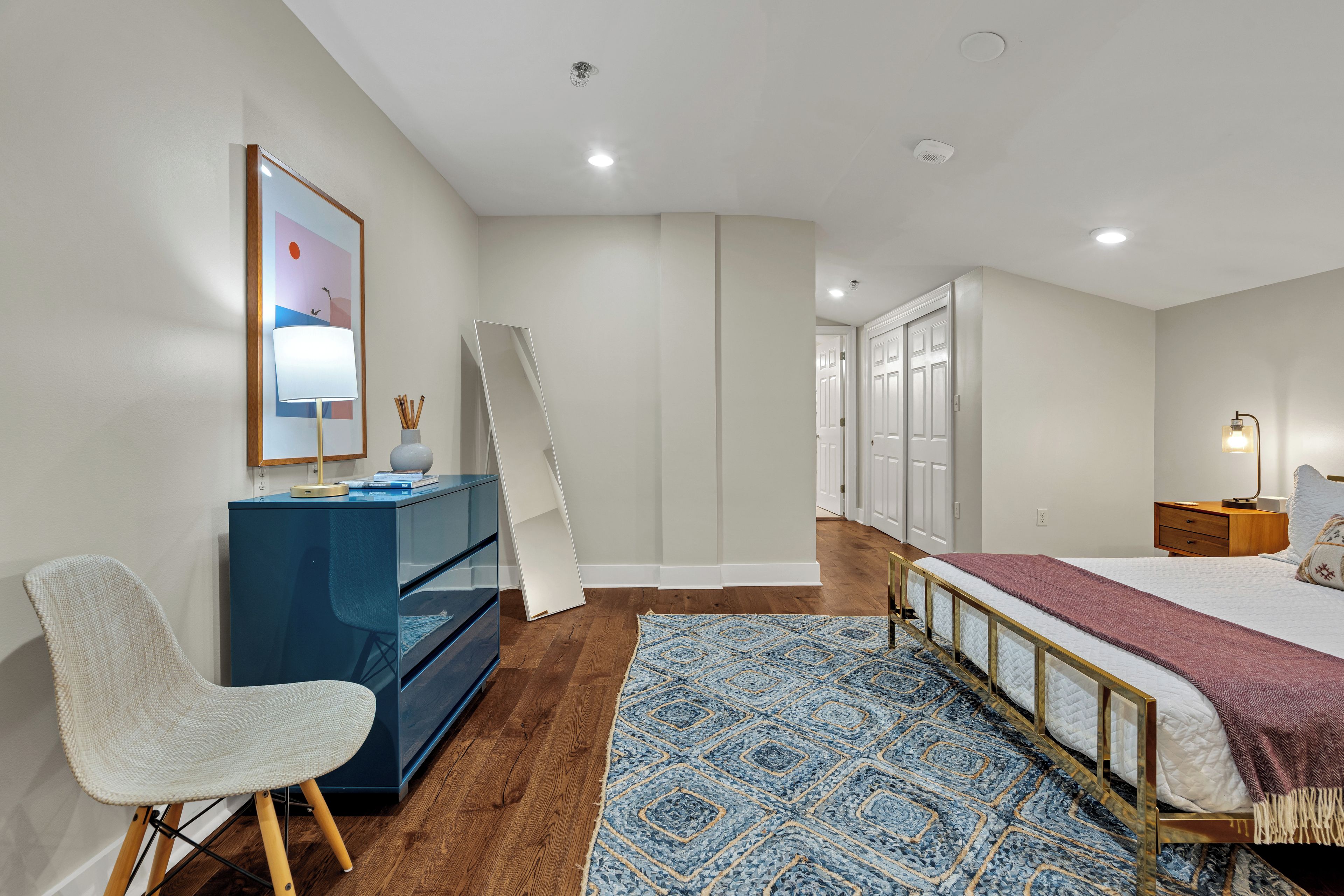 A bedroom with a bed, a desk with a chair, a dresser, a rug, and a door leading to another room.