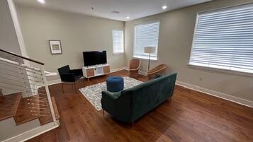 A spacious living room with hardwood flooring, a green sofa, a flat-screen TV, and a staircase.