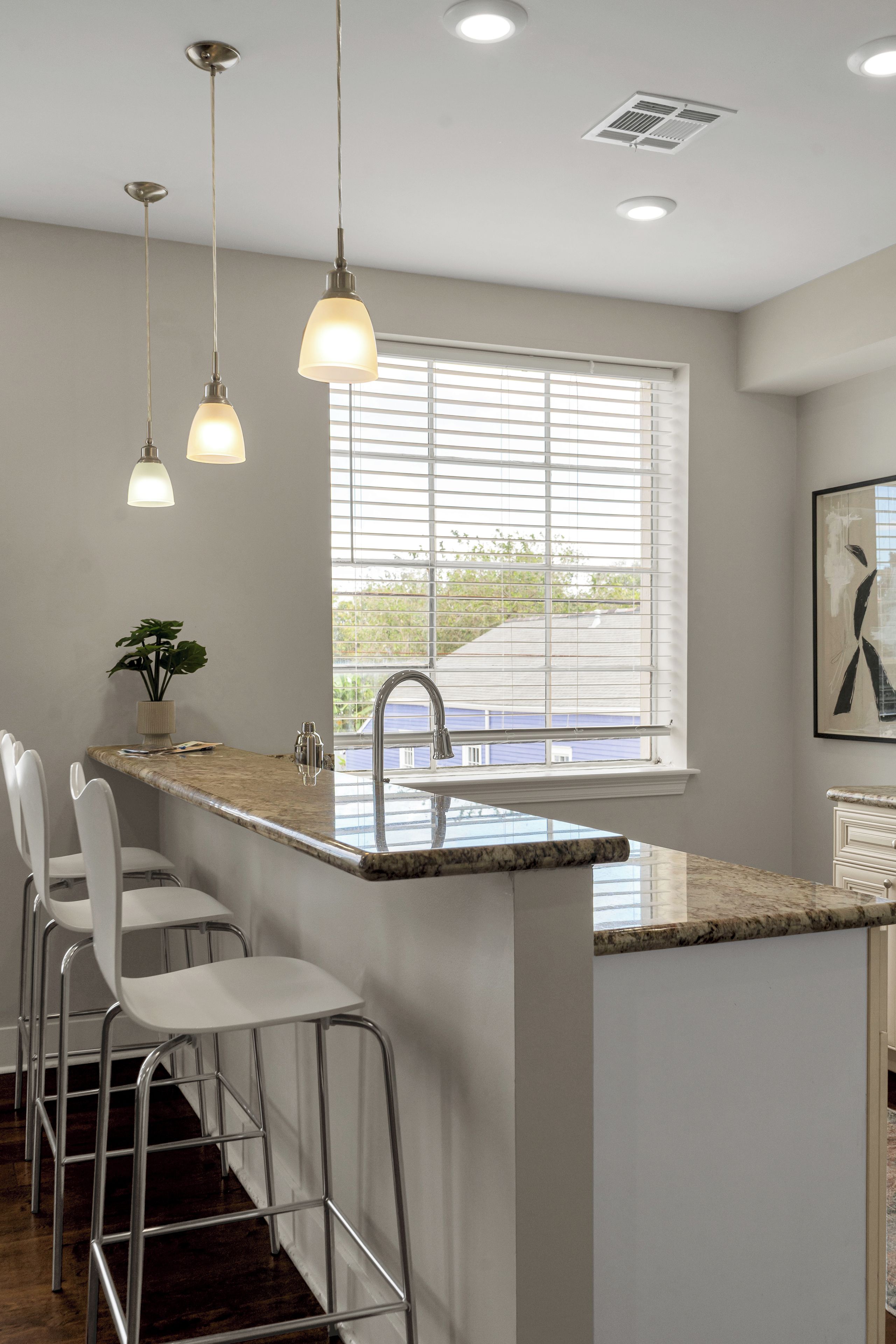 A kitchen with a marble countertop, stainless steel bar stools, pendant lighting, and a window with blinds.