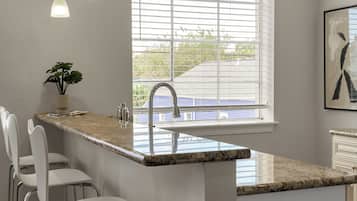A kitchen with a marble countertop, stainless steel bar stools, pendant lighting, and a window with blinds.