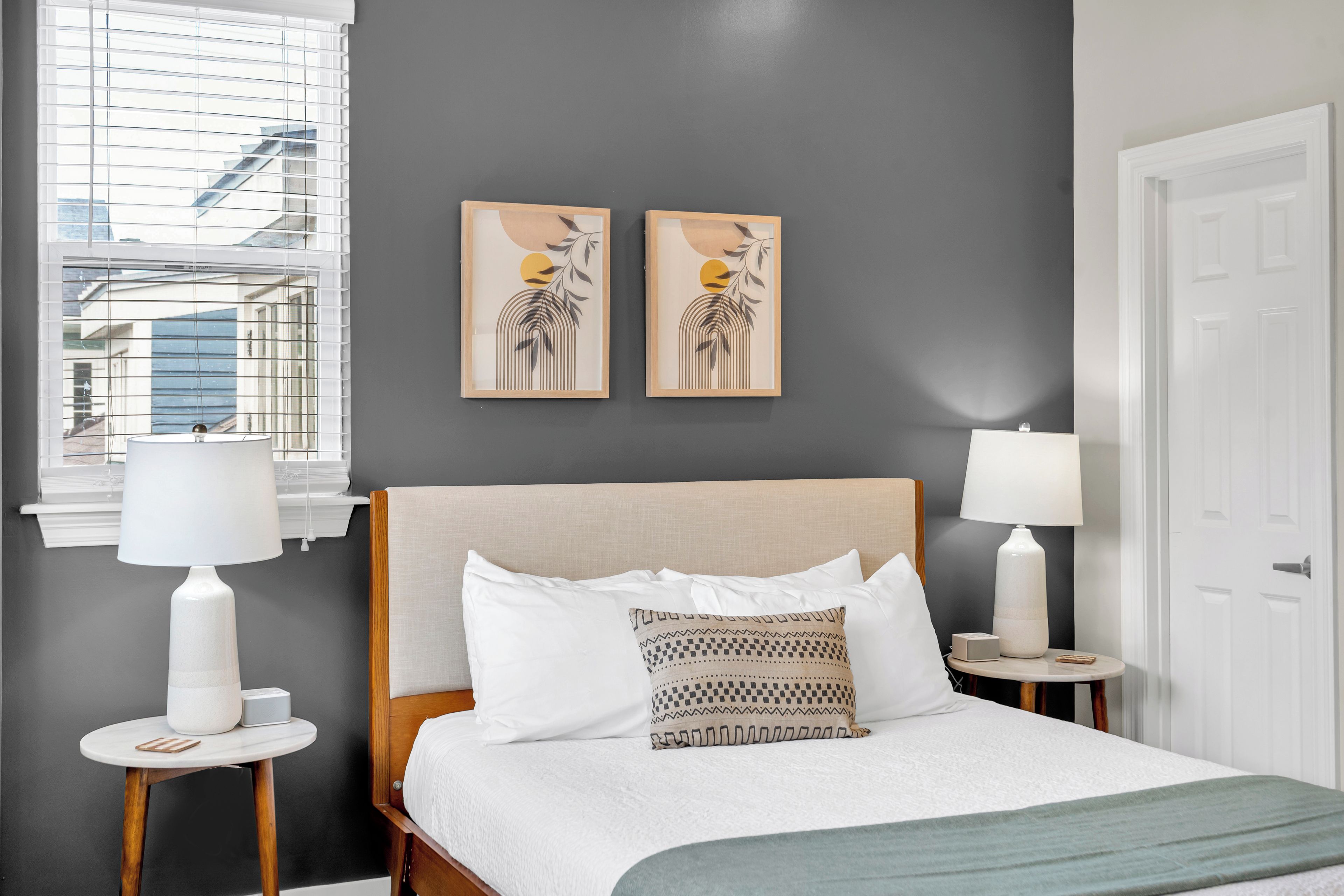 A hotel room with a bed, two bedside tables with lamps, a window with blinds, and two wall art pieces.