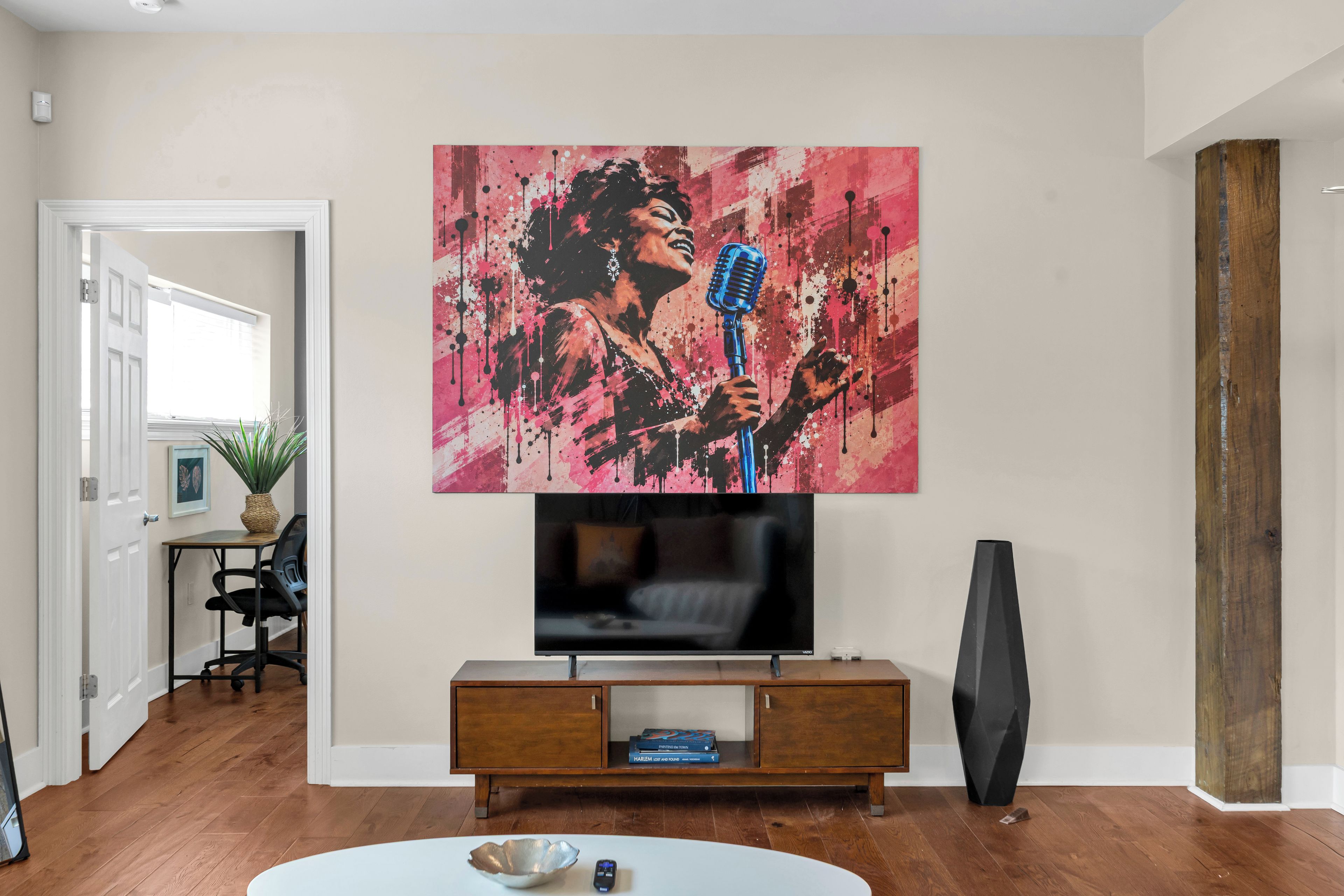 A living room with a flat-screen TV on a wooden stand, a painting of a person with a microphone, and a modern black sculpture.