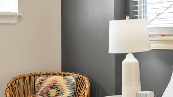 Ein Zimmer mit einem Bett, einem Korbstuhl mit gemustertem Kissen, einer weißen Lampe auf einem kleinen Tisch und einem Fenster mit Jalousien.