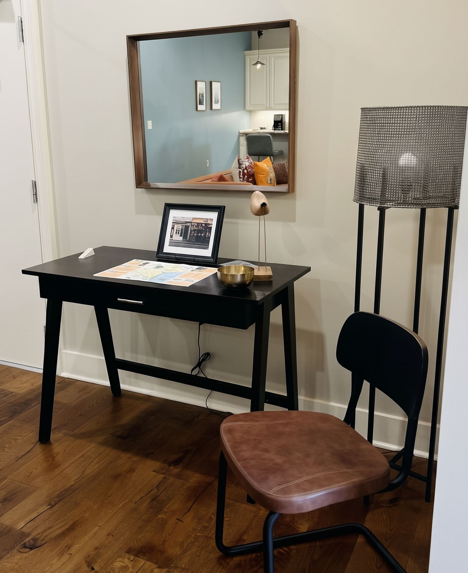 Ein Zimmer mit Schreibtisch, Stuhl und einem kleinen Hocker. An der Wand hängt ein Spiegel und auf dem Schreibtisch steht eine Lampe.
