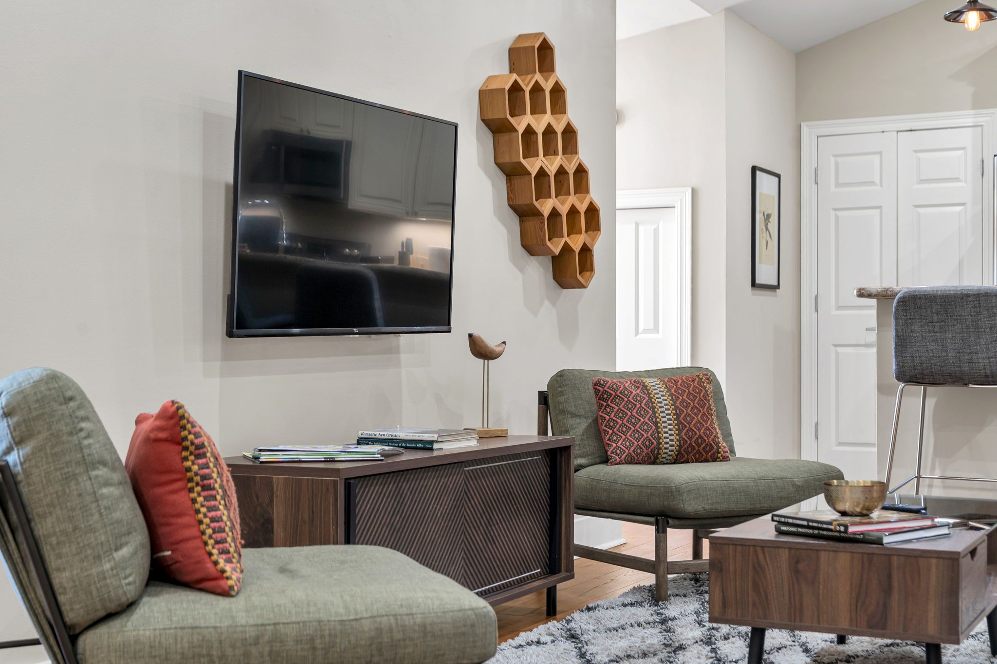 A modern living room with a flat-screen TV, two green armchairs, a wooden coffee table, and a decorative wooden shelf.