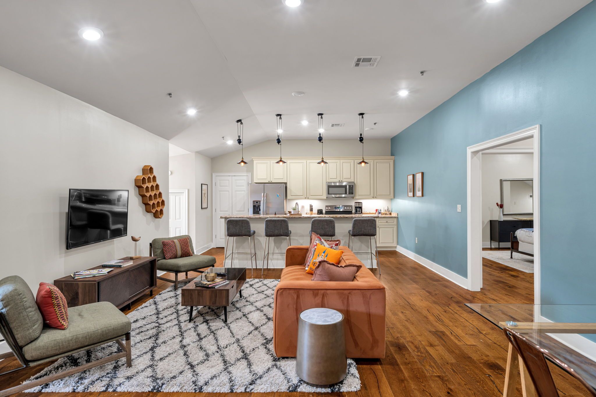 A modern living room with a sofa, coffee table, and TV. There is a kitchen with cabinets and appliances in the background.