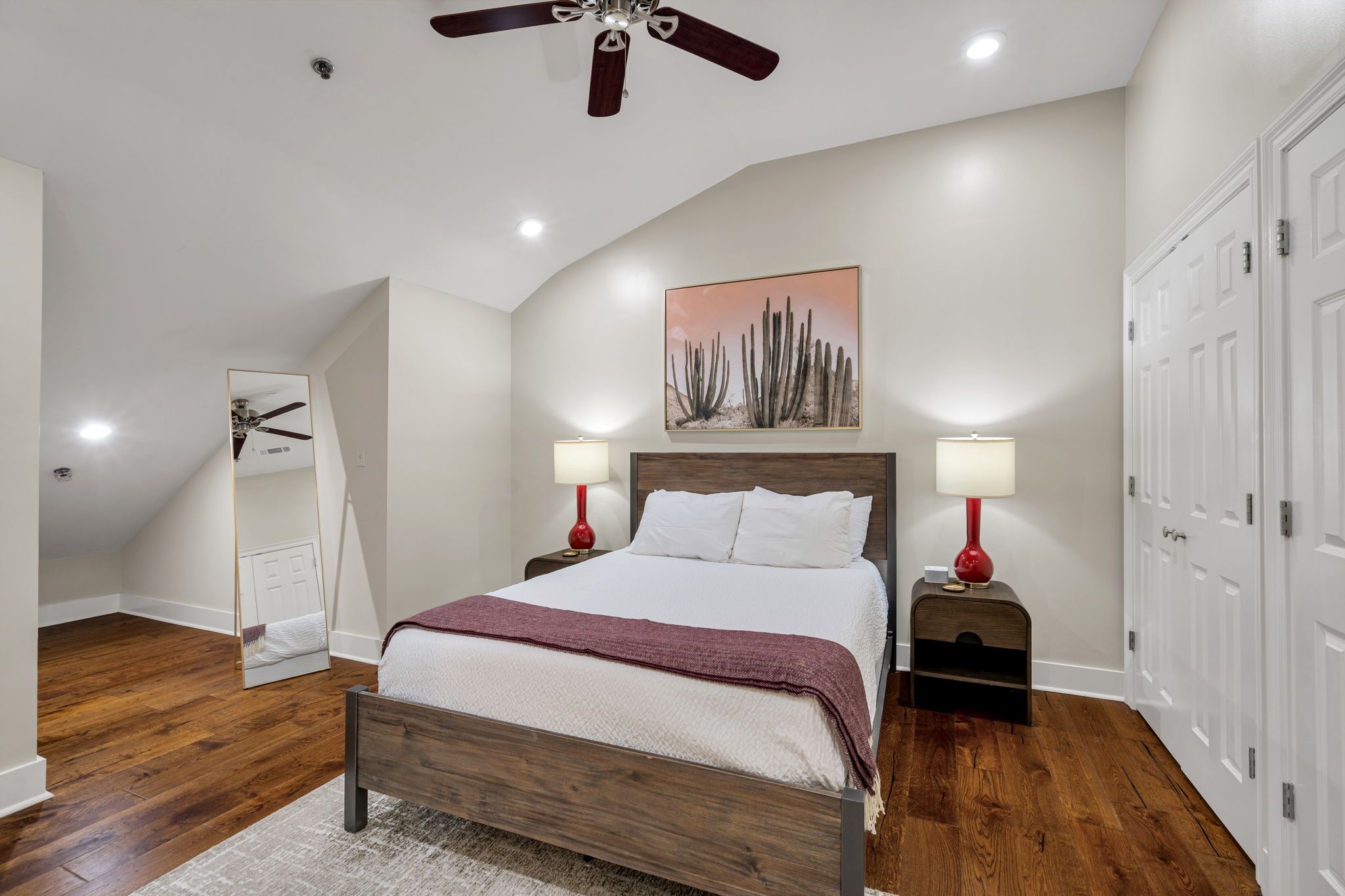 A bedroom with a bed, two bedside tables with lamps, a ceiling fan, a painting on the wall, and a door.
