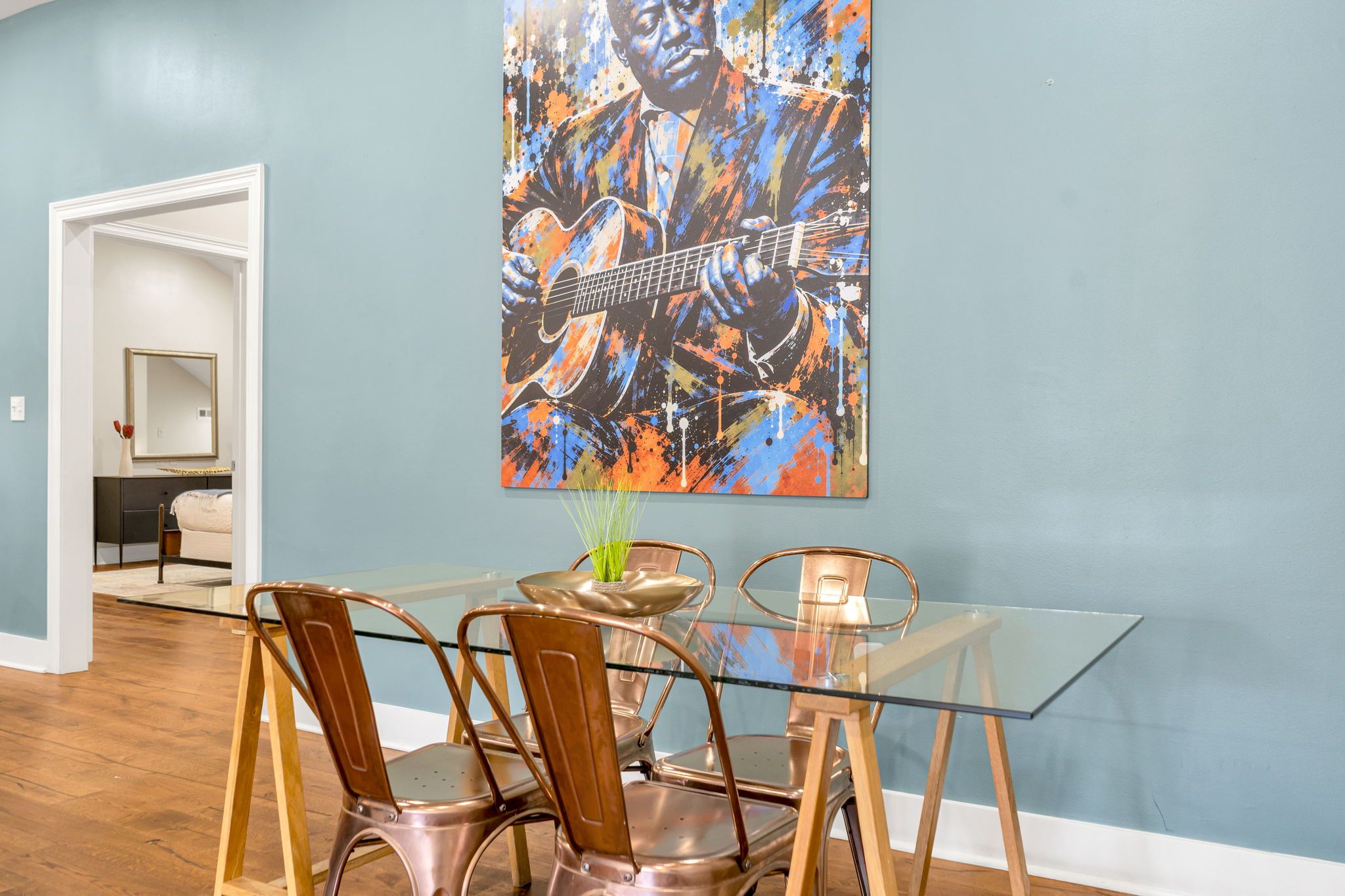 A dining area with a glass table and metal chairs, a painting on the wall, and a hallway leading to another room.