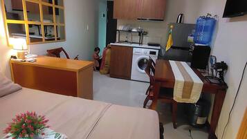 A compact hotel room with a bed, a desk, a washing machine, and a kitchenette.