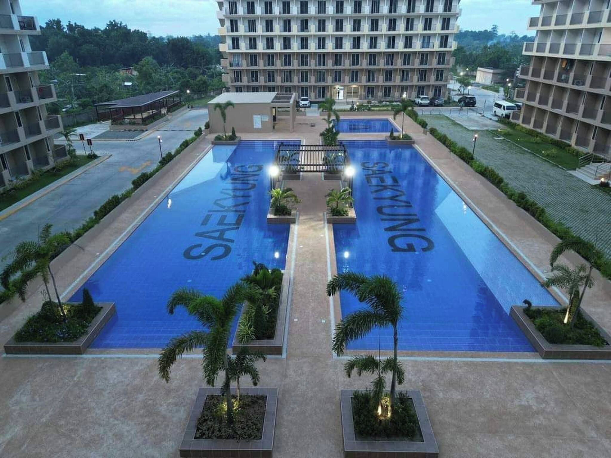 Una piscina con la marca 'SAEKYUNG', rodeada de jardines con palmeras y un edificio de varios pisos al fondo.