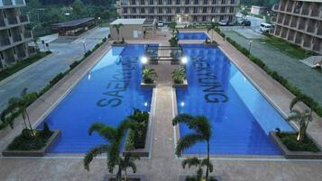 Una piscina con la marca 'SAEKYUNG', rodeada de jardines con palmeras y un edificio de varios pisos al fondo.