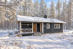 Une cabane dotée d’un porche, d’une porte et d’une cheminée, entourée d’une forêt enneigée.