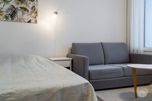Una habitación moderna con una cama, un sofá gris, una mesita blanca pequeña y un aplique de pared.
