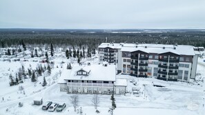 A multi-story building with a flat roof, surrounded by snow-covered ground and trees, indicating a winter setting.