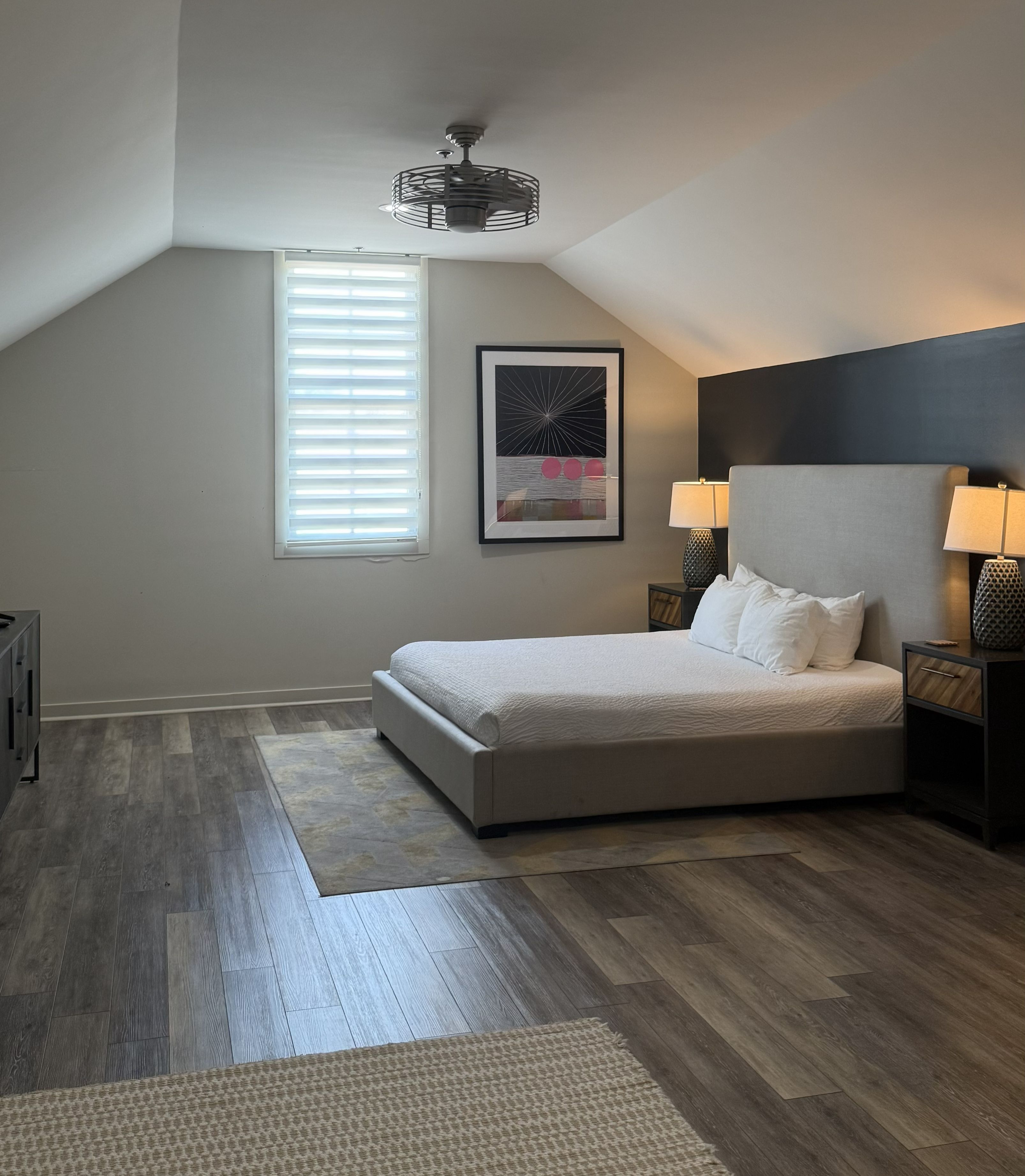 A bedroom with a bed, a window with blinds, a ceiling fan, and a nightstand with a lamp.