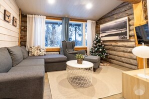 A log cabin interior with a gray sofa, armchair, and a small table with a plant. A Christmas tree is visible in the corner. - Villa Quality side B by Holiday in Lapland (Ruka)