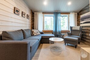Ein gemütliches Wohnzimmer mit einer Couch, einem Sessel und einem Couchtisch, Holzwänden und großen Fenstern mit Vorhängen.