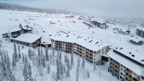 A snow-covered resort with multiple buildings, including a hotel with balconies and a parking area.