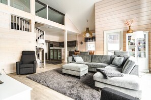 Ein modernes Wohnzimmer mit einem grauen Ecksofa, einem schwarzen Sessel und einem weißen Couchtisch. Eine Treppe führt in eine obere Ebene, und im Hintergrund ist eine Küche zu sehen.