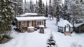 Una cabana de fusta amb un teulat cobert de neu, una llar de pedra i un cobert petit en un bosc nevat.