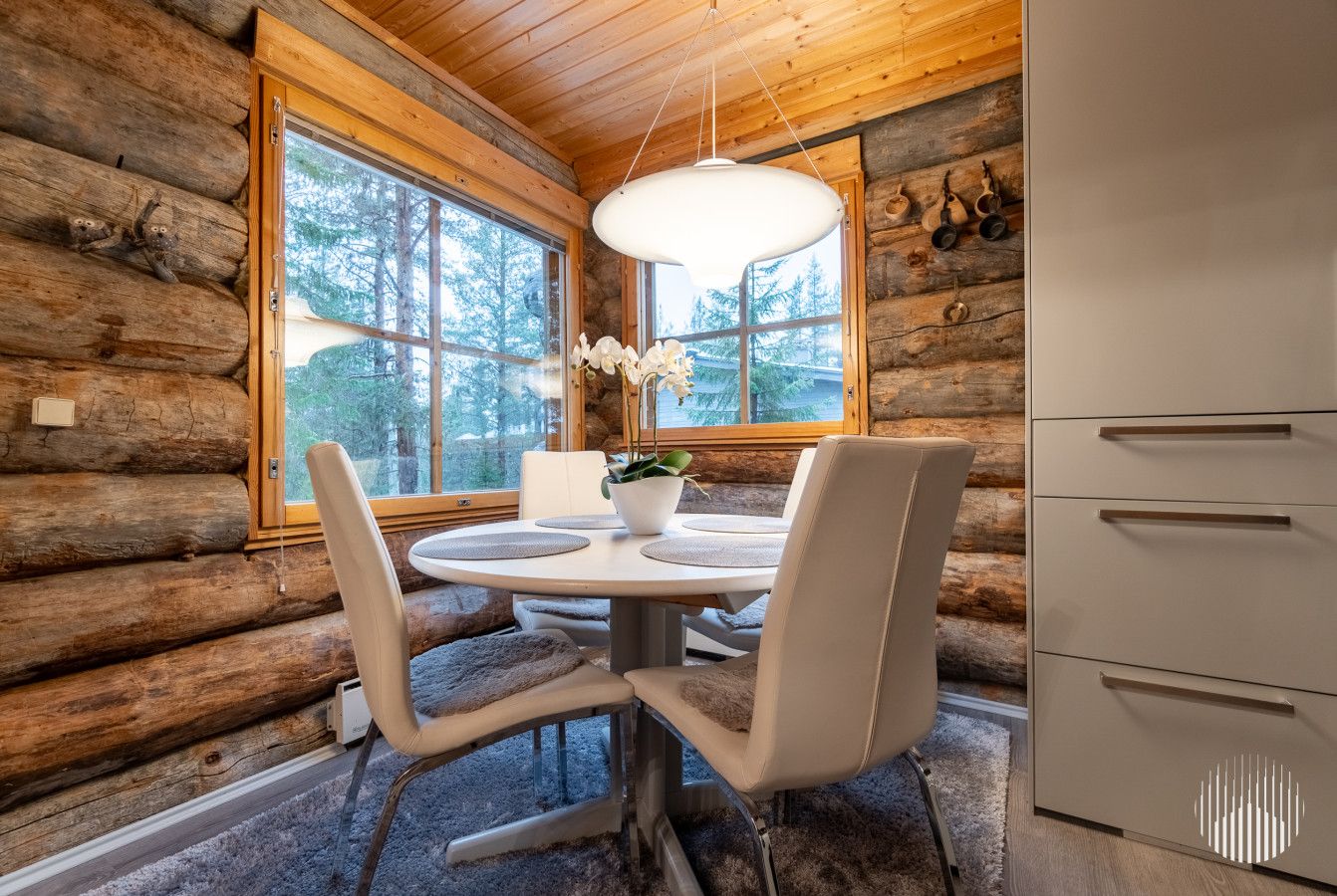 En stuga byggd av timmer med en inredning som inkluderar ett matbord, stolar, en hängande lampa och ett köksområde.