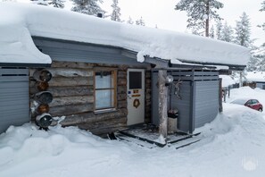 Une cabane en rondins dont le toit est recouvert de neige, avec une porte ornée d’une guirlande et un petit porche.