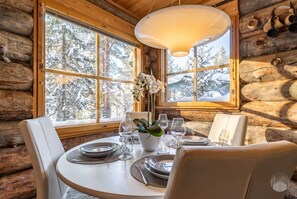 A log cabin interior with a dining table set for four, surrounded by chairs, and large windows offering a view of snow-covered trees. - HilleVilla by Holiday in Lapland (Ylläs)